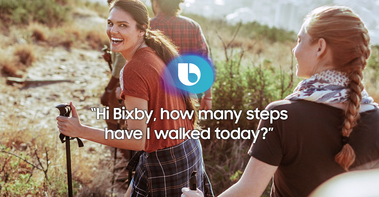 An image of displaying the message "Hi Bixby, how many steps have I walked today?" on a picture of a woman laughing while hiking with friends