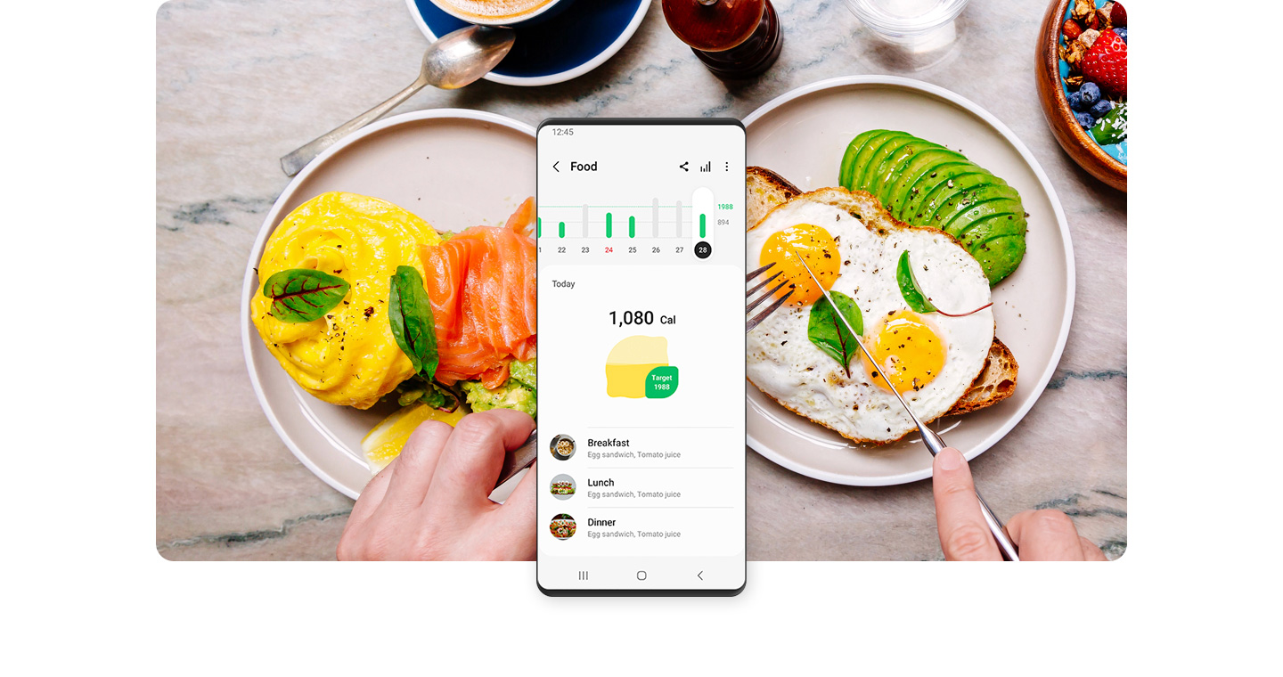Galaxy smartphone is shown displaying the day's nutrition calorie count. Behind the smartphone, a breakfast containing eggs, toast and avocado is shown surrounded by a berry cake, coffee and a plate with salmon, eggs benedict and avocado toast.