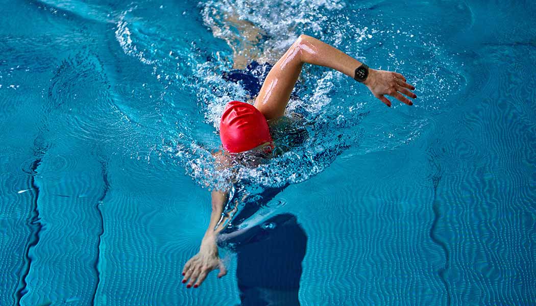 Icons of different exercises can be seen and the swimming icon is highlighted. A woman is swimming in a swimming pool while wearing a Galaxy Watch4 device.