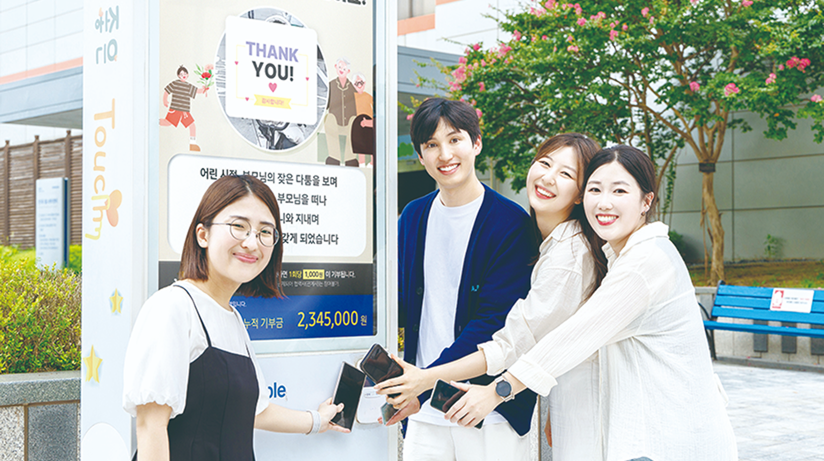 네 명의 사람들이 나눔키오스크 화면 앞에 나란히 서서 휴대폰을 터치하며 기부에 참여하는 모습