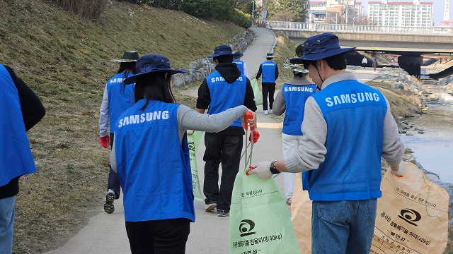 Samsung Electronics employees, wearing blue vests and hats, walk along a stream path carrying tongs and trash bags as they engage in a cleanup activity.
