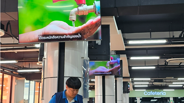 A water-saving campaign image emphasizing the importance of water is displayed on monitors inside a company cafeteria.