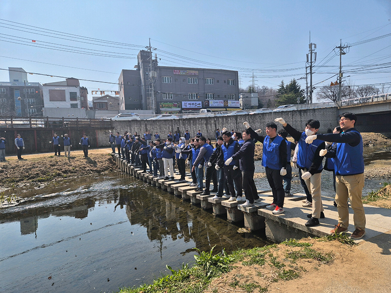 Samsung Electronics employees, wearing blue vests and gloves, stand in a line along a walkway above the stream, ready to begin a cleanup activity.