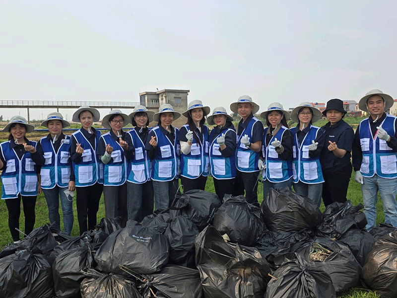 Samsung Electronics employees, wearing blue vests and hats, stand in a line in front of collected garbage bags, posing for a group photo after a cleanup activity.